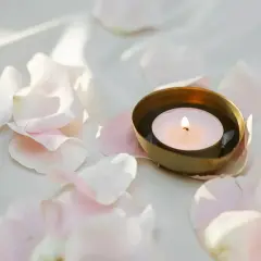 Delikatny płomień tealight pali się w złotej miseczce, otoczony przez jasnoróżowe płatki róż.