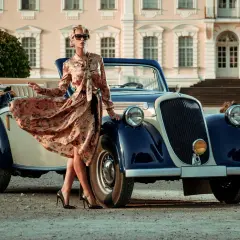 Elegancka kobieta w kwiecistej sukience i okularach przeciwsłonecznych pozuje przy zabytkowym kabriolecie. Stylowe lata 20. w pełnej krasie.