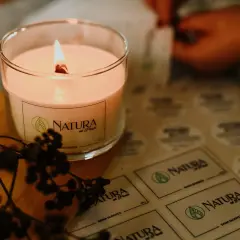 Płonąca, naturalna świeca zapachowa w szkle, obok gałązka z owocami. W tle arkusz z etykietami "NATURA od Pauli" - inspiracja, jak zrobić naturalne świece zapachowe.