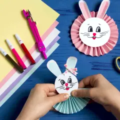 Ręce składają wielkanocnego zajączka z papieru, obok kolorowy papier, pisaki i nożyczki.