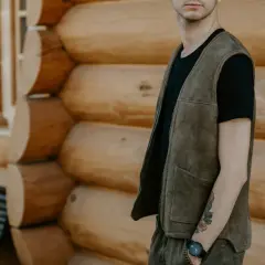 Mężczyzna w zamszowym serdaku i czarnej koszulce stoi na tle drewnianego domu.