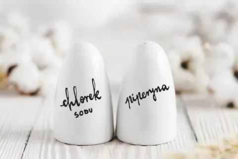 Białe solniczki z napisem "chlorek sodu" i "piperyna" – idealny pomysł, co kupić na parapetówkę, by dodać kuchni stylu.