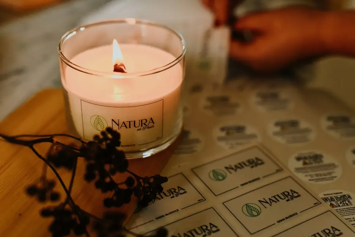 Płonąca, naturalna świeca zapachowa w szkle, obok gałązka z owocami. W tle arkusz z etykietami "NATURA od Pauli" - inspiracja, jak zrobić naturalne świece zapachowe.