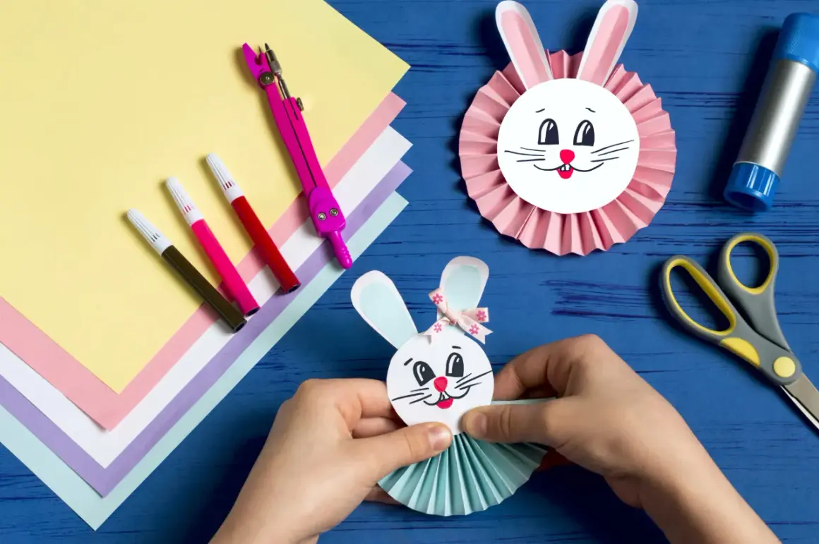 Ręce składają wielkanocnego zajączka z papieru, obok kolorowy papier, pisaki i nożyczki.