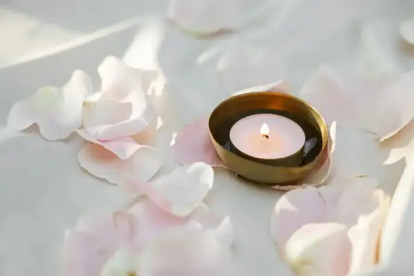 Delikatny płomień tealight pali się w złotej miseczce, otoczony przez jasnoróżowe płatki róż.