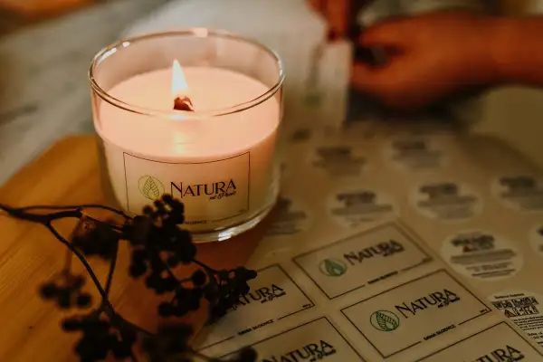 Płonąca, naturalna świeca zapachowa w szkle, obok gałązka z owocami. W tle arkusz z etykietami "NATURA od Pauli" - inspiracja, jak zrobić naturalne świece zapachowe.