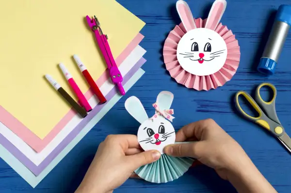 Ręce składają wielkanocnego zajączka z papieru, obok kolorowy papier, pisaki i nożyczki.